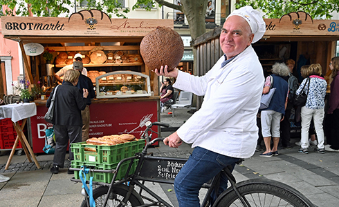 baeckerei-neulinger-brotmarkt2024-LudwigNeulinger-490x300 Unser Chef, Bäckermeister Ludwig Neulinger, auf seinem Fahrrad hält einen großen Laib Brot vor dem Stand der Bäckerei Neulinger auf dem Brotmarkt hoch.